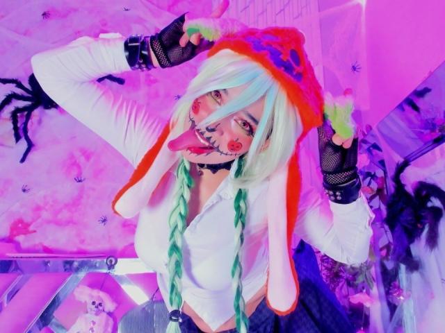 KiraBerry with green pigtails, white shirt, rainbow hat, black fishnet gloves, tongue out, face paint, and large tits, in a pink-lit room, holding hat.