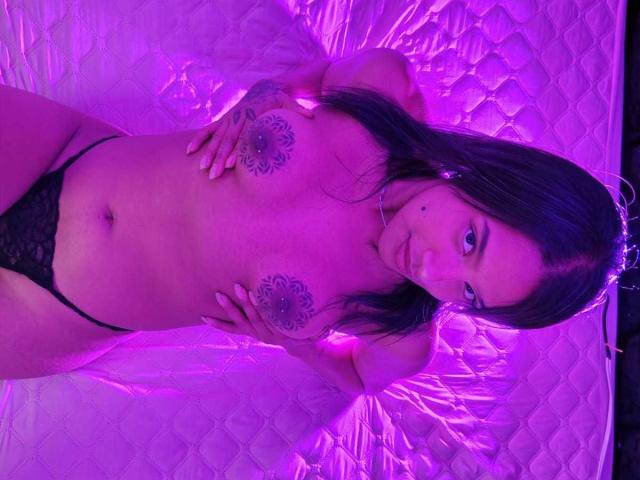 AmberPieri with dark hair, tattoos on her tits, black lace panties, hands on her chest, purple lighting, standing on a white quilted mattress.