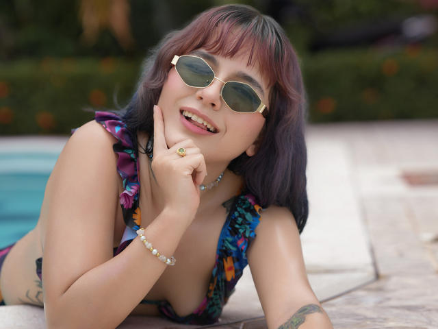 HairyValeri lying by the pool, wearing a colorful floral bikini, sunglasses, and a bead bracelet, with her finger on her chin, smiling at the camera. Her tits are visible.