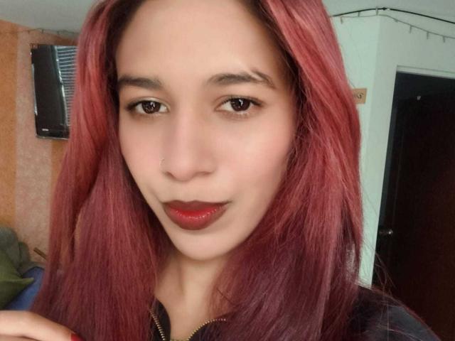 ValerieWoods has red, straight hair, brown eyes, and red lipstick, with a slight smirk. She's wearing a black and yellow plaid shirt. Her nose is pierced.