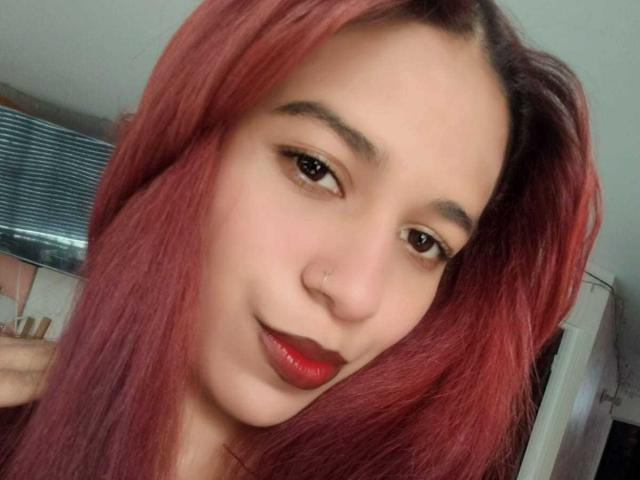 ValerieWoods has red hair, a nose ring, and red lipstick. She's looking at the camera with a slight smile. Her brown eyes are half-open. Her tits are not visible.
