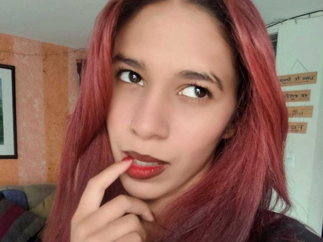 ValerieWoods with red hair, brown eyes, and red lipstick, biting her finger, showing glossy red lips, and a small nose ring. Her hand is near her mouth.