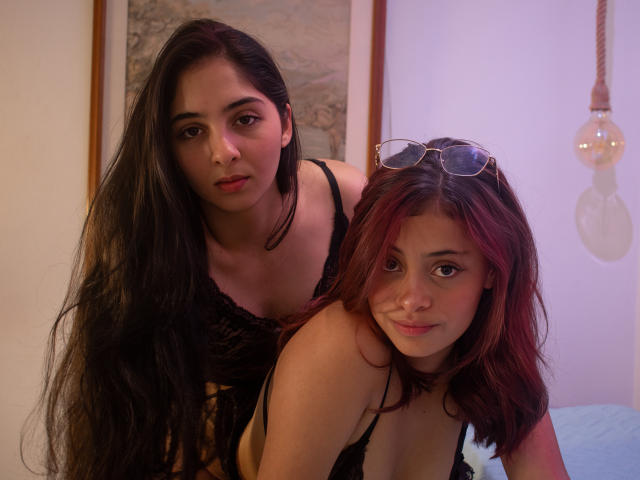 EvaAndEmma with long black hair and EvaAndEmma with red hair, both wearing black lace tops, standing close, EvaAndEmma's tits visible, red-haired EvaAndEmma has glasses on head.