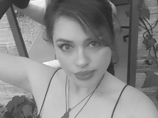 Autoportrait en noir et blanc de PaulettWatsoon, avec du rouge à lèvres foncé, un bras levé et une coiffure désordonnée, portant un haut à bretelles fines et un collier, montrant sa poitrine et un regard confiant..