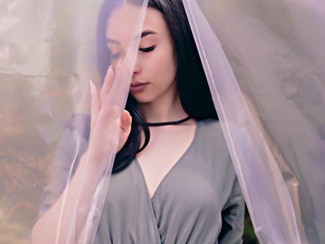 CamilleRios with pale skin, long black hair, and a grey V-neck dress, gently holding a translucent curtain, her eyes closed, and black eyeliner visible, touching her cheek.