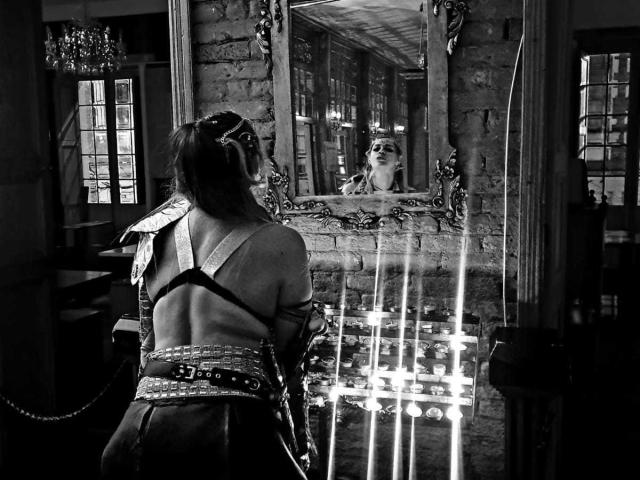 Fotografia in bianco e nero di PaulettWatsoon, con la schiena e le braccia scoperte, che indossa una camicetta e una cintura in stile giapponese, in piedi di fronte a uno specchio che riflette il suo viso..