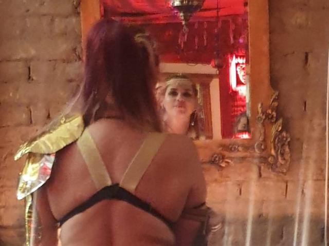 PaulettWatsoon avec des cheveux violets, une armure dorée sur les épaules, un bikini noir à bretelles, regardant un miroir, un arrière-plan rouge, le reflet montrant un visage confiant, légèrement flou..