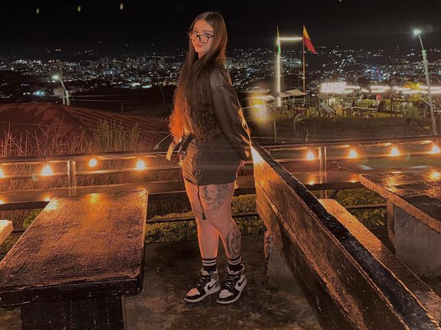 MileyBaker with long brown hair, glasses, furry black sweater, black shorts, tattoos on legs, black-and-white sneakers, standing on wooden bench, string lights, night, city skyline.