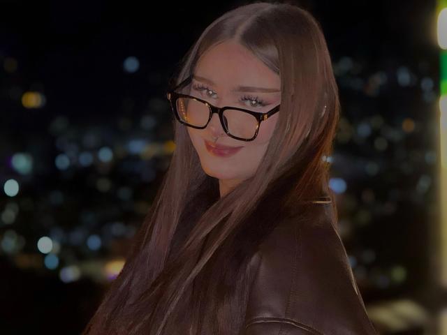 MileyBaker with long brown hair, wearing black tortoiseshell glasses, brown leather jacket, and a slight smile, in a dark, bokeh city night background.