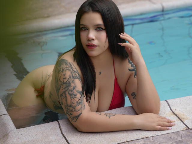 AryButty with dark hair, red bikini, tattoos on arms and chest, leaning in pool, looking at camera, hand on head, blue water, pale skin, large boobs, sexy.