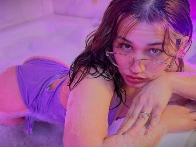 ElodieGardner with wet brown hair, wearing glasses and purple lingerie, leans over in a bath, her ass up, looking at the camera with a pouty expression, ring on her hand.