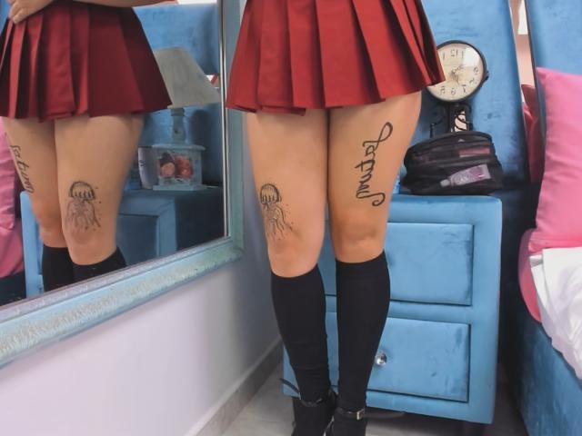 Genesiss's legs, wearing a red skirt, black knee socks, and black shoes, with a jellyfish tattoo on the right thigh and "Angel" tattooed on the left.