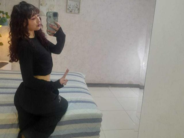 LilyDickenson with curly brown hair, black long-sleeve crop top, yellow belt, and black skirt, taking a mirror selfie, showing her curvy ass and holding a pink dildo.