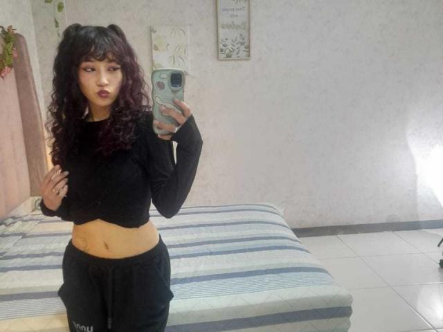 LilyDickenson with curly black hair, wearing a black crop top and black pants, taking a selfie showing her midriff and black lipstick, standing in a room with a striped bed.
