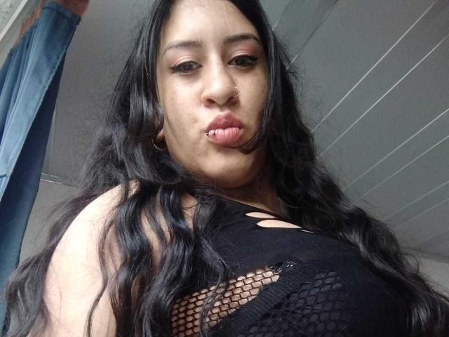 AmbeerKiss tiene el pelo largo y negro, un piercing en la nariz y está frunciendo el ceño con los labios ligeramente entreabiertos. Lleva una camiseta de malla negra que deja ver sus grandes senos..