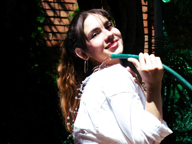 LindaPerez with wavy brown hair, white shirt, holding green hose, smiling, red lipstick, hoop earrings, sunlit, tree in background, water droplets on shirt.