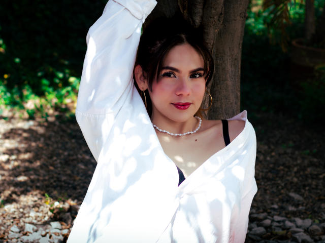 LindaPerez with dark hair, red lipstick, white shirt, black bra, pearl necklace, arm raised, leaning against tree, looking at camera, sunlight filtering through leaves.