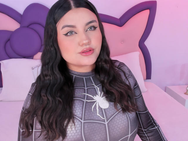 MarlaBriard with long black wavy hair, wearing a spiderweb-patterned black latex top, showing her tits, has a nose ring, and pink lipstick, sitting on a bed.