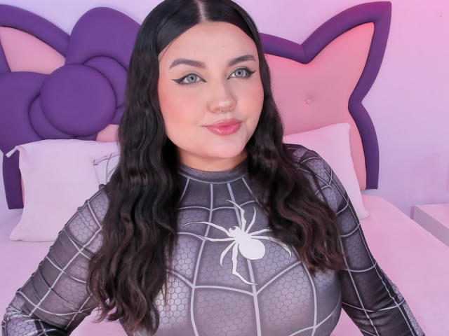 MarlaBriard with long black wavy hair, wearing a spider web-patterned, grey, tight, latex top, showing her big, round, shiny tits, standing in front of a purple, star-shaped headboard.