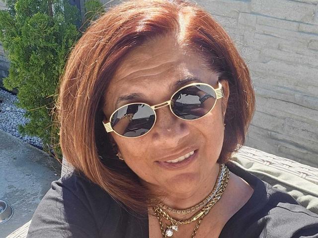 Eine elegante, aber freche Frau mit roten Haaren, die goldene Sonnenbrillen und mehrere Halsketten trägt und in die Kamera lächelt. Ihre Brüste sind teilweise unter einem schwarzen Oberteil sichtbar. Die Aufnahme entstand im Freien bei Sonnenschein..