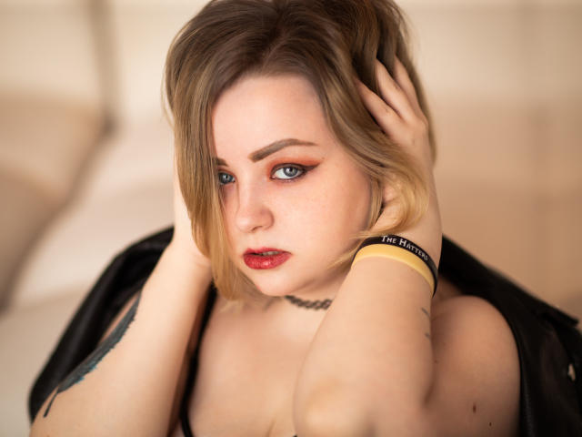 MissEmmar, avec ses cheveux blonds, son rouge à lèvres rouge, et ses yeux bleus, les mains dans les cheveux, un collier noir, un bracelet jaune, le regard baissé, un visage légèrement constellé de taches de rousseur, une lumière douce, un tatouage sur le bras gauche..