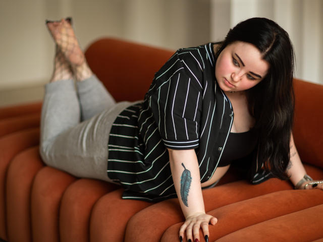 MissEmmar è sdraiata su un divano arancione, indossa una camicia a righe nere e pantaloni grigi. Ha un tatuaggio a forma di piuma sul braccio destro e calze a rete nere..