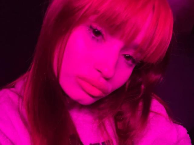 HenryDellarocco with pink-tinted red hair, pouty lips, and a slight tilt of the head, wearing a hoodie, in a pink-lit selfie.