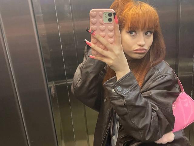 HenryDellaracco with red hair, wearing a brown leather jacket, holding a pink phone with heart patterns, in an elevator, looking serious, pink bag on shoulder, long nails.