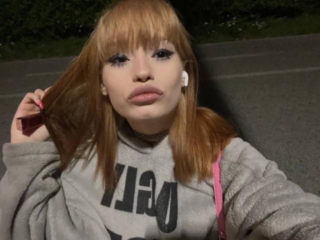 HenryDellarocco with light brown hair, wearing a gray hoodie, holding hair with right hand, pink strap, white earbuds, pouty lips, and dark eye makeup. Nighttime, street.