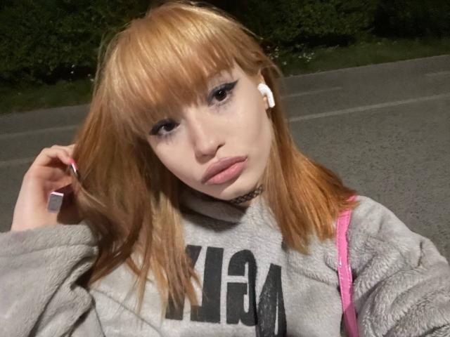 HenryDellarocco a les cheveux bruns clairs, porte un sweat à capuche gris, une bandoulière de sac à dos rose, des écouteurs blancs et un collier noir. Il a les mains dans les cheveux, regarde la caméra et a les lèvres roses..