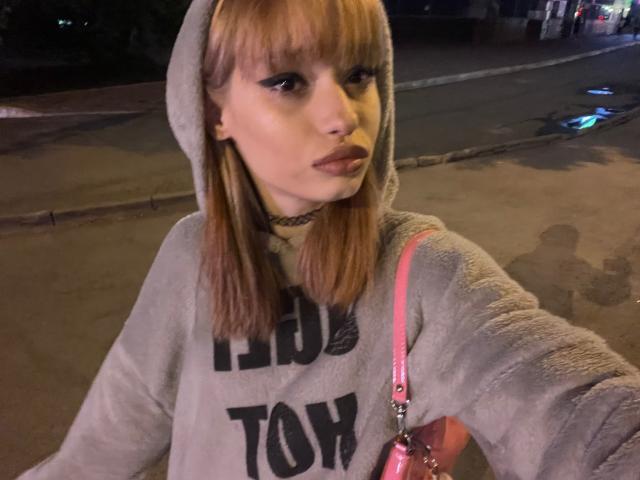HenryDellarocco with blonde bangs, wearing a gray hoodie, pink crossbody, and black choker, looking into the camera at night, with dark eyeliner and pink lipstick.