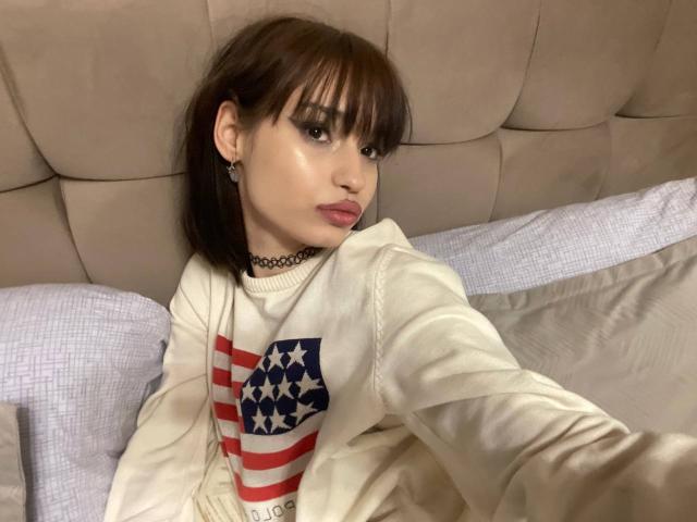 HenryDellarocco with brown pigtails, wearing a white sweatshirt with an American flag design, sitting on a bed, choker necklace, and looking at the camera with a slight smirk.