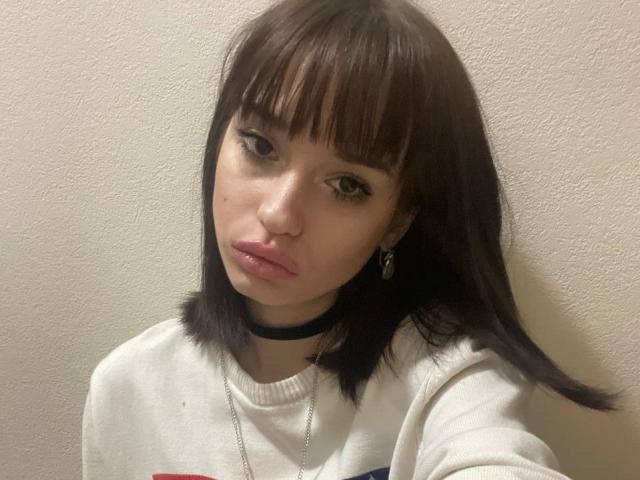 HenryDellarocco a les cheveux bruns avec une frange, un pull blanc, un collier noir et des boucles d'oreilles brillantes, avec un visage boudeur et un maquillage discret. Elle porte un collier en argent..