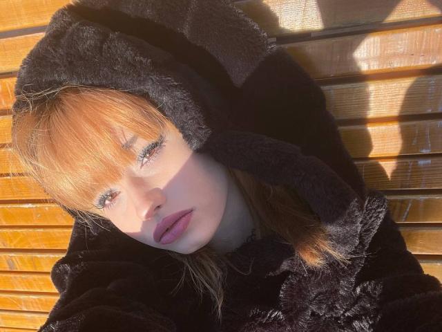 HenryDellarocco with orange hair, blue eyes, and purple lipstick, wearing a black fuzzy hood, looking up, sunlight casting shadows on wooden slats.
