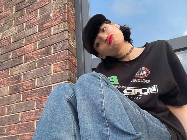 Henry Dellarocco, avec ses cheveux noirs courts, sa casquette noire, son rouge à lèvres rouge, son t-shirt noir, son jean bleu et son collier, faisant la moue, assis contre un mur de briques, regardant vers le haut..