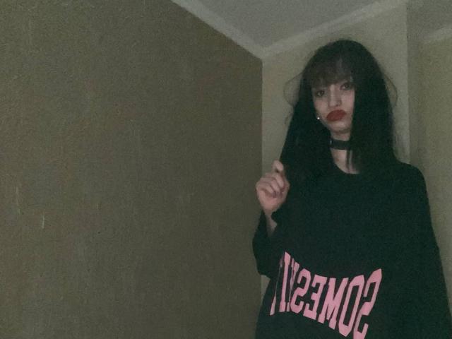 HenryDellarocco with long black hair, dark lipstick, wearing a black choker and oversized black shirt with pink text, standing in dimly lit room, raising right hand.