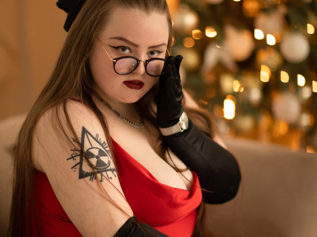 MissEmmar with glasses, red dress, black gloves, and long brown hair, showing a tattoo on her arm, in a cozy, warmly lit setting, touching her glasses.