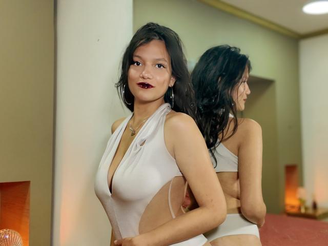 Melody, met golvend zwart haar, donkerrode lipstick en een witte top met uitsparingen die haar borsten en billen laat zien, staat voor een spiegel en glimlacht licht..