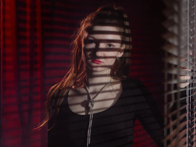 NikaFlorence with long brown hair, black top, and silver necklace, standing in shadows with striped blinds casting patterns on her face and chest. Red light highlights her pink lips.