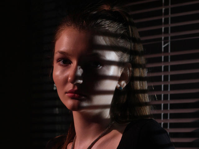 NikaFlorence has shiny, sweaty skin with light shadows from blinds across her face, brown hair, and blue earrings, wearing a black top, looking serious with pink lips.