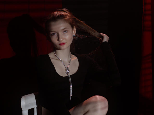 NikaFlorence with fair skin, red lipstick, black top, and long brown hair, holding it with one hand, wearing a silver necklace, sitting on a white chair in dark.