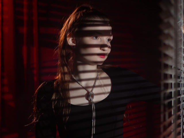 NikaFlorence with long brown ponytail, black top, silver necklace, red lipstick, shadowed by window blinds, looking to the side, red light in background.