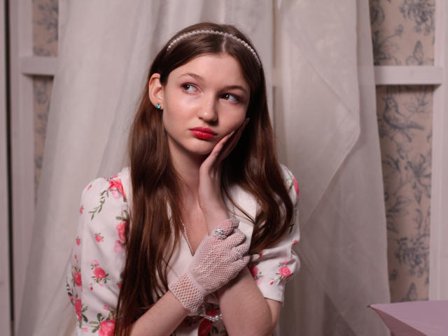NikaFlorence with long brown hair, pink floral dress, white lace glove, red lipstick, looking to the side, hand on cheek, soft expression, pearl headband, blue earrings, indoors.