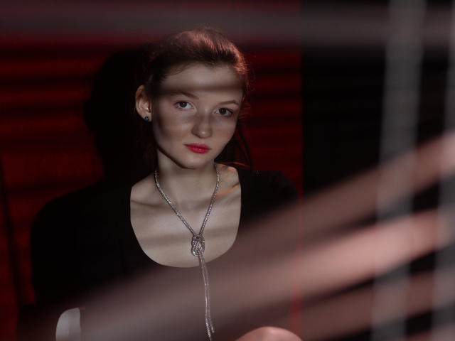 NikaFlorence with red lipstick, wearing a black top and silver necklace, has light skin and brown hair, looking slightly to the side, with blinds in the foreground.
