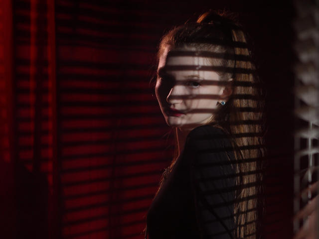 NikaFlorence's face, shadowed by blinds, shows her pale skin and subtle smile. Her long brown hair is partially lit, and she wears a black top, looking over her shoulder.