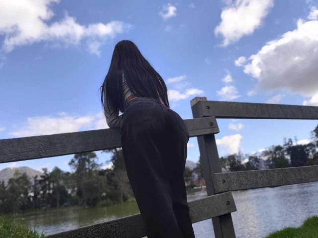 GinaRider leaning over a wooden fence, butt in tight black pants, long black hair, blue sky with clouds, water and greenery in background, visible butt and lower back.