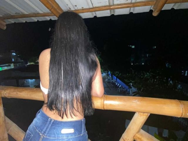 GinaRider with long black hair, facing away, in a white top, showing her ass in blue jeans, leaning on a bamboo railing at night.