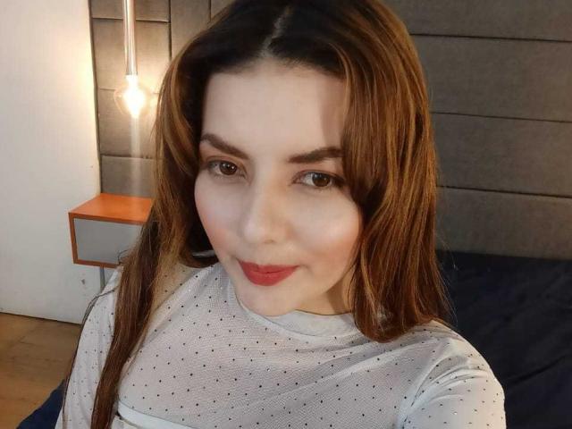 MandyMouses ha lunghi capelli castani, occhi castani e rossetto rosso. Indossa una maglietta bianca con pois e sorride alla fotocamera. Il suo seno è piccolo ed è visibile nella foto..