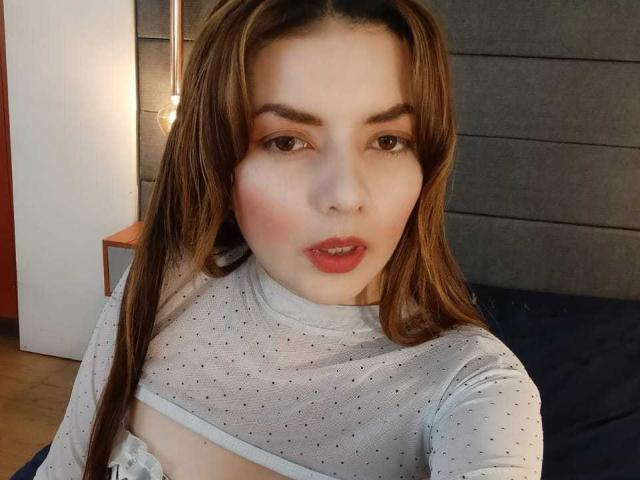 MandyMouses con capelli castani, pelle chiara e labbra rosa, che indossa una maglietta bianca trasparente a pois, che mette in mostra il suo seno piccolo, e guarda la fotocamera con un'espressione di bocca leggermente aperta..