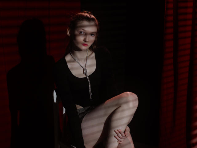 NikaFlorence with light skin, red lipstick, black long-sleeve top, and shorts, sitting with one leg up, shadow on wall, silver necklace, red-striped light.